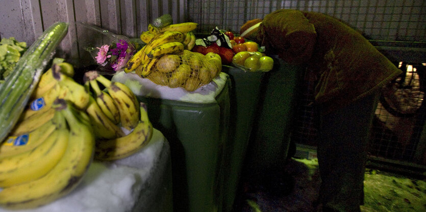 Bananen und Kartoffeln, Gurken liegen auf schneebedekcten Mülltonnen. Dahinter sucht ein Mensch nach weiterem Essen