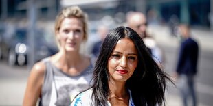 Sawsan Chebli ist zu sehen, sie blickt leicht an der Kamera vorbei und trägt die Haare offen, hinter ihr steht eine Frau, der Kamera-Fokus liegt auf Chebli