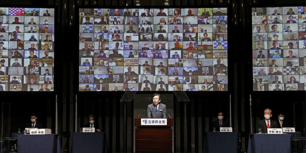 Ein Mann steht an einem Rednerpult, hinter im ein großer Bildschirm mit zugeschalteten Teilnehmer*innen