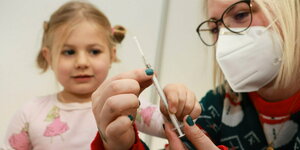 Ein fünfjähriges Mädchen lässt sich vor ihrer ersten Corona-­Impfung von einer Impf­assistentin eine Spritze zeigen