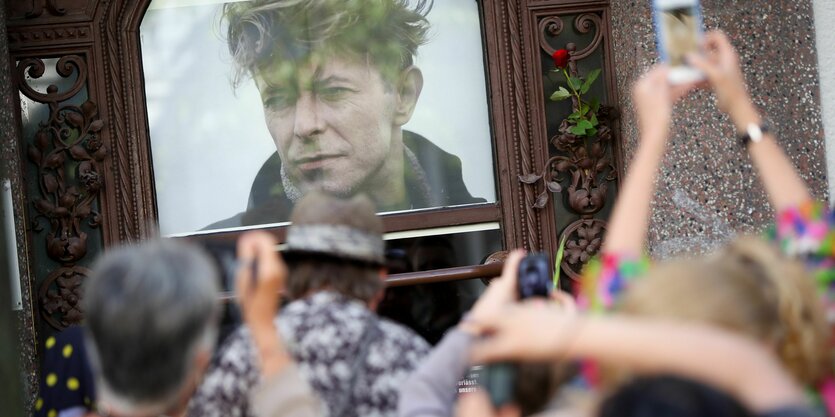 Zahlreiche Menschen stehen 2016 in Berlin in der Hauptstraße 155 vor einem Porträtfoto von David Bowie