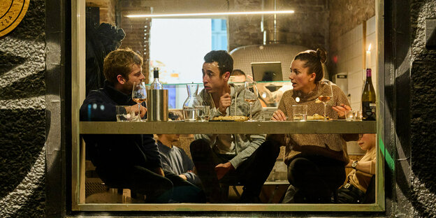 Zwei Männer und einen Frau Sitzen in einer Pizzeria auf einer Fesnterbank mit Wein, Gläser und Teller vor sich.