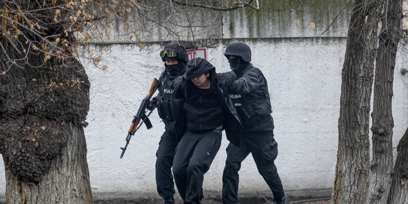 Zwei bewaffnete Polizisten in Uniform führen einen Zivilisten ab