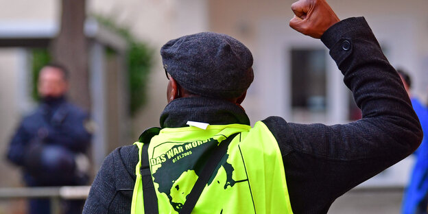 Ein Mann (Ansicht von Hinten) mit gelber Oury-Jalloh-Warnweste streckt seine Faust in Richtung Polizei