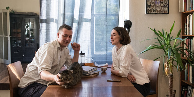 Ein Mann und eine Frau sitzen sich am Tisch gegenüber; er krautl die Katze, die auf dem Tisch hockt
