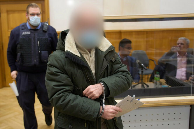 Ein Mann mit verpixeltem Gesicht in Handschellen, hinter ihm steht ein Polizist