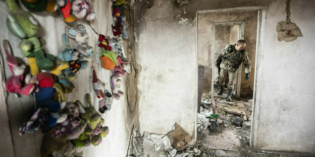 Bunte Kuscheltiere hängen an der Wand eines zerstörten, verlassenen Zimmers, in einem Türrahmen ist ein Soldat zu sehen