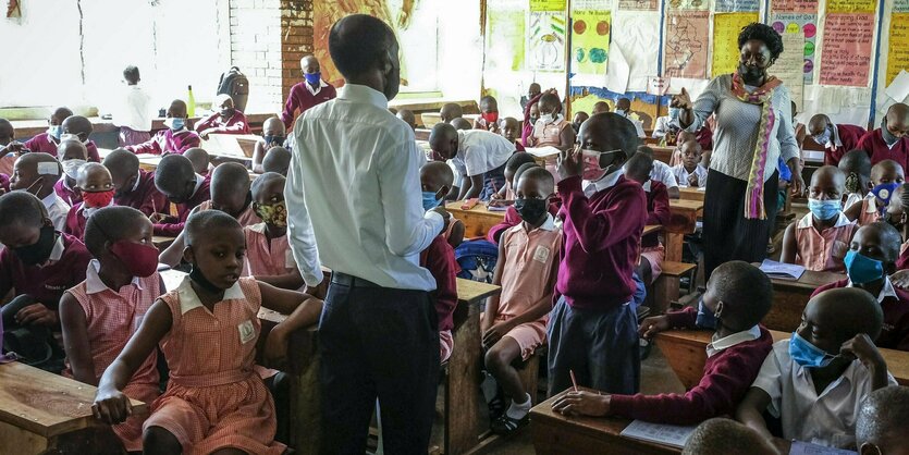 Eine Lehrer und eine Lehrerin stehen in einer Klasse mit vielen Kindern, die Mundschutz tragen