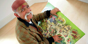 Ein älterer Mann mit roter Mütze und grauem Schnurrbart hält ein Kinderbuch, von oben fotografiert