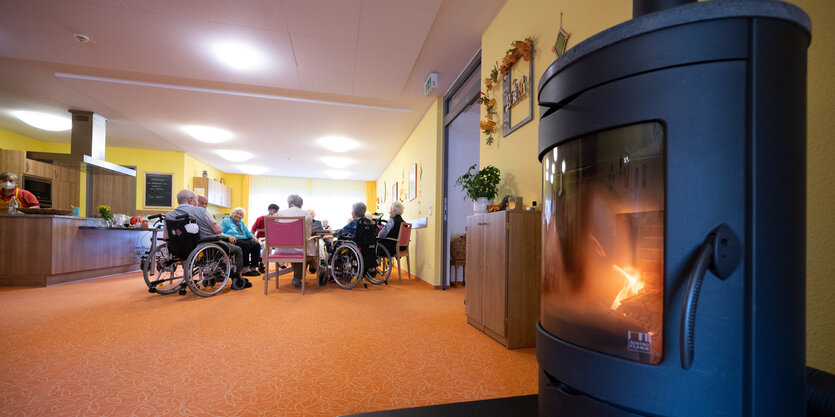 Menschen sitzen im Altenheim an einem Tisch. Im Vordergrund brennt ein Feuer in einem Kaminofen.