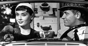 Szene aus "Sabrina": eine Frau und ein Mann sitzen in einem Cabriolet, die Frau (links) hält einen Pudel auf dem Schoß