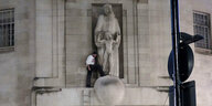 Ein Mann steht vor einer Skulptur hoch am Haus der BBC. die skupltur : ein bärtiger Mann hält einen nackten Jungen zwischen seinen Armen