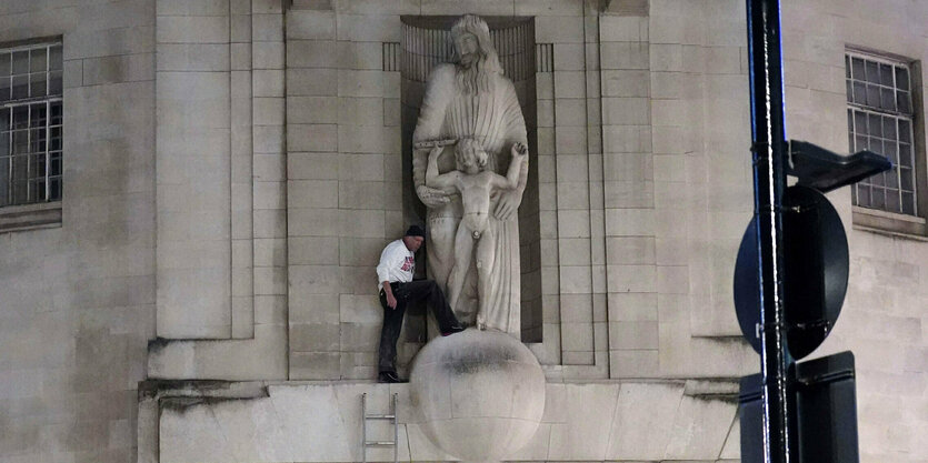 Ein Mann steht vor einer Skulptur hoch am Haus der BBC. die skupltur : ein bärtiger Mann hält einen nackten Jungen zwischen seinen Armen