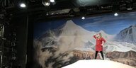 Die Autorin Stefanie Sargnagel steht auf einer Bühne auf einem riesigen Bauch, im Hintergrund sind Berge zu sehen.