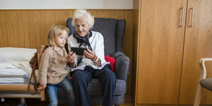 Eine Seniorin sitzt auf einem Sessel neben einem einzelbett und zeigt einem kleinem Mädchen etwas auf einem Smartphone