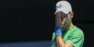 Novak Ðoković, ein Tennisspieler. Er trägt ein grünes T-Shirt und eine graue Cap. Mit der Hand fasst er sich an die Stirn.