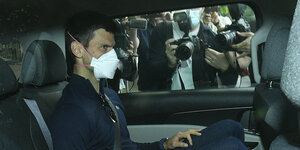 Tennisstar Novak Djokovic auf dem Rücksitz von einem Auto. Er trägt eine Maske. Hinter dem Fenster stehen Fotograf*innen und versuchen Bilder von ihm zu machen.