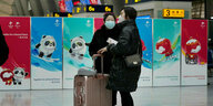 Zwei Frauen mit Koffern und Mundschutz stehen vor Olympia-Plakaten