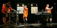 Drei Menschen in orangener Kleidung auf einer Bühne