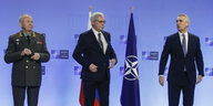 NATO Generalsekretär Stoltenberg steht mit zwei russischen Ministern auf einem Podium, zwischen ihnen die Fahren der NATO, hinter Stoltenberg die Fahne Russlands