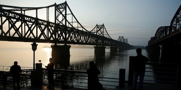 Menschen stehen im Abendlicht vor einer Brücke über einen Fluss