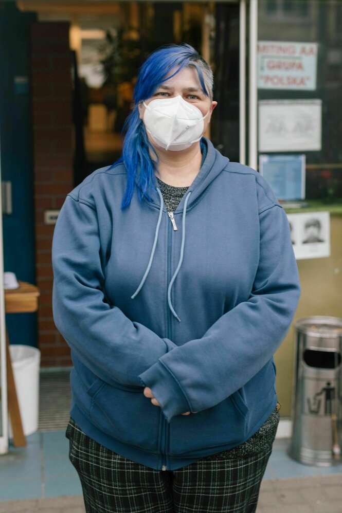 Eine Frau mit blau gefärbten langen Haaren und blauer Kapuzenjacke steht vor einem Eingang