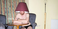 Ein Mann sitzt in einer Wohnung und trägt einen Lampenschirm auf dem Kopf