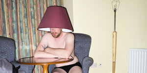 Ein Mann sitzt in einer Wohnung und trägt einen Lampenschirm auf dem Kopf