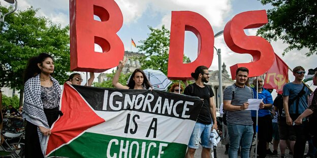 Eine Frau steht mit einer Flagge mit der Schrift "Ignorance is a choice" vor einer Gruppe bei einem Protest. Im Hintegrund sind große Buchstaben zu sehen: BDS