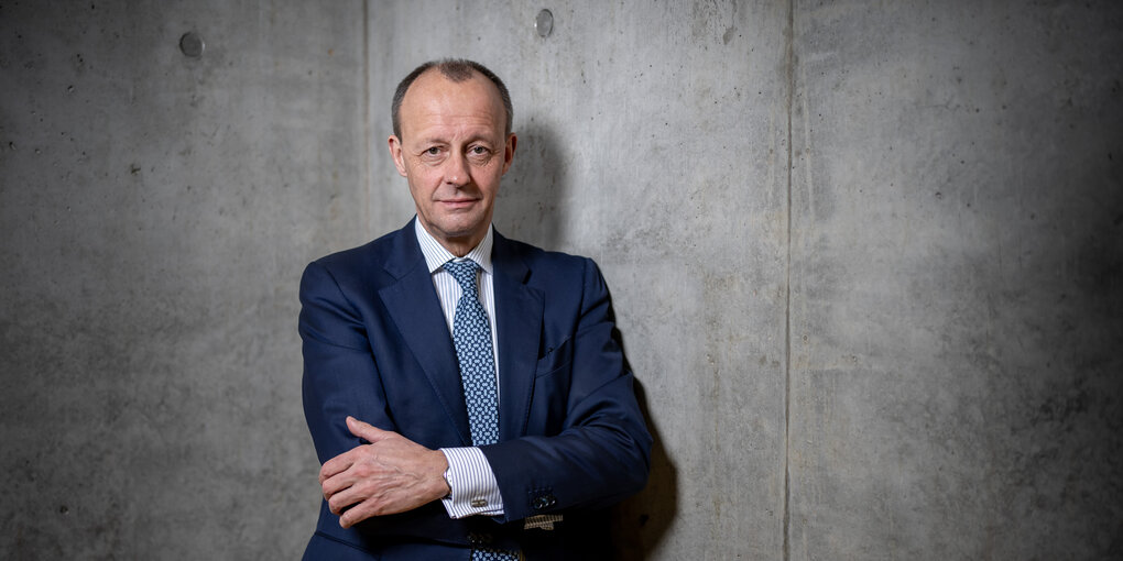 Friedrich Merz im Anzug und mit verschränkten Armen vor einer Betonwand