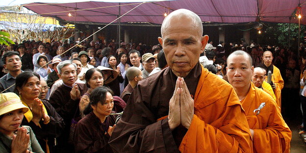 Thich Nhat Hanh im Mönchsgewand und betender Haltung mit dem Rücken zu einer Menschenmenge vor einer Pagode in Vietnam