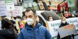 Cem Özdemir mit Mund/Nasenschutz. Im Hintergrund demonstrierende Bauern mit Plakaten und Traktoren
