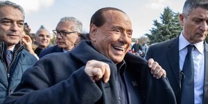 Italiens Präsident Berlusconi lacht und hält das Revers seines Mantels. Um ihn herum stehen Männer in Anzügen
