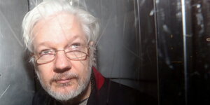 Julian Assange hat weiße Haare und eine Brille und schaut schrräg nach oben