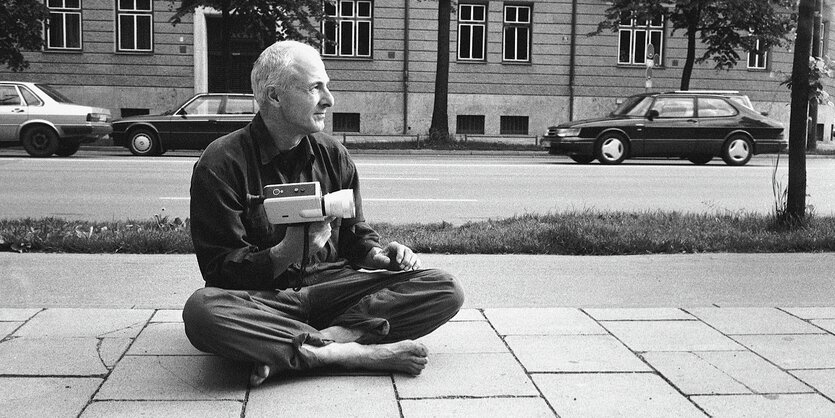 Herbert Achternbusch sitzt im Schneidersitz auf dem Pflaster mit einer Kamera