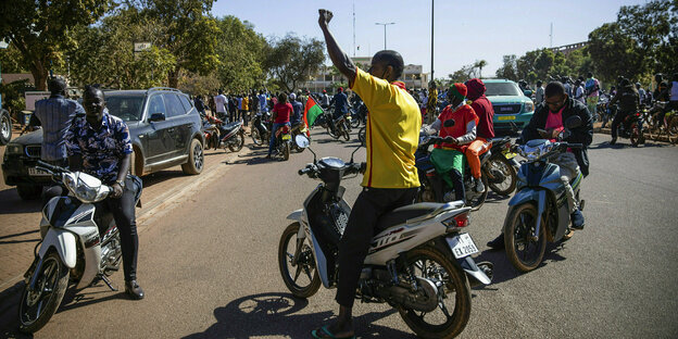 Männer auf Motorrädern stehen auf einer Straße, einer reckt die Faust, im Hintergrund eine demonstrierende Menschenmenge