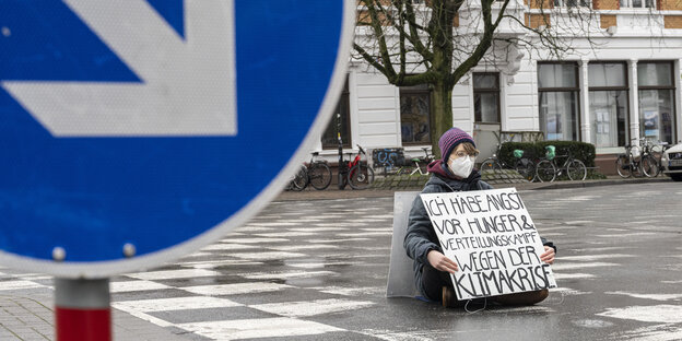 Eine Aktivistin sitzt auf einer Straße und hält ein Schild mit der Aufschrift: „Ich habe Angst vor Hunger und Verteilungskampf wegen der Klimakrise“