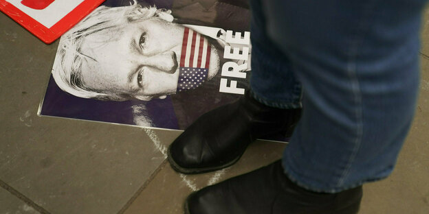 Schwarze Lederschuhe stehen neben einem Plakat mit dem Gesicht Assanges und der amerikanischen Flagge über seinem Mund