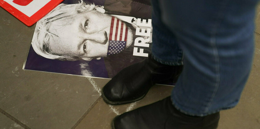 Schwarze Lederschuhe stehen neben einem Plakat mit dem Gesicht Assanges und der amerikanischen Flagge über seinem Mund