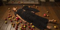 Eine Frau in einem schwarzen Kleid liegt zwischen Äpfeln auf dem Fußboden vor einem Kamin