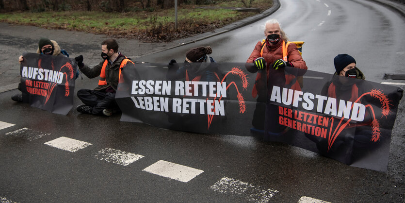 Fünf Menschen unterschiedlicher Altersgruppen sitzen in Winterkleidung und Warnwesten auf einer kleinen asphaltierten Straße und halten Transparente, auf denen "Essen retter, Leben retten" steht.