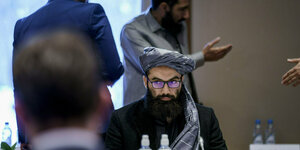 Ein Taliban sitzt am Tisch, vor ihm ein Mannim anzug,hinter ihm gestikulierende Hände