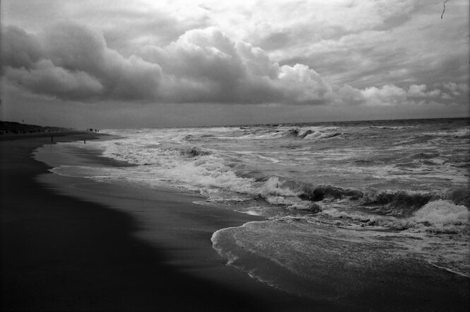 Die Aufnahme in Schwarzweiß zeigt dunkle Wolken, den Strand und stürmische See