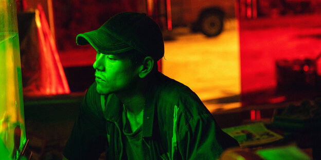 Xueming (Eddie Peng), von einem Aquarium grün angeleuchtet, sitzt vor rotem und gelbem Hintergrund.