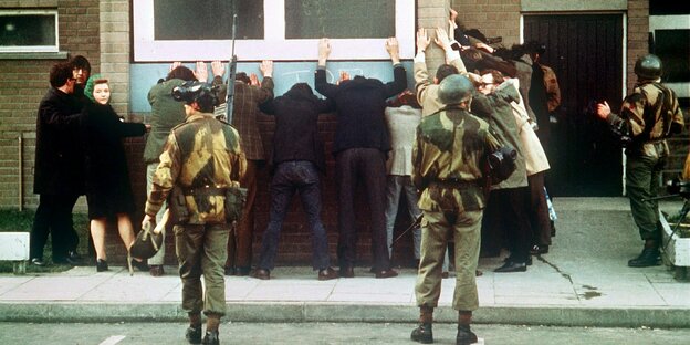 Am Bloody Sunday: An einer Wand stehen Menschen mit erhobenen Händen aufgereiht, hinter ihnen britische Soldaten.