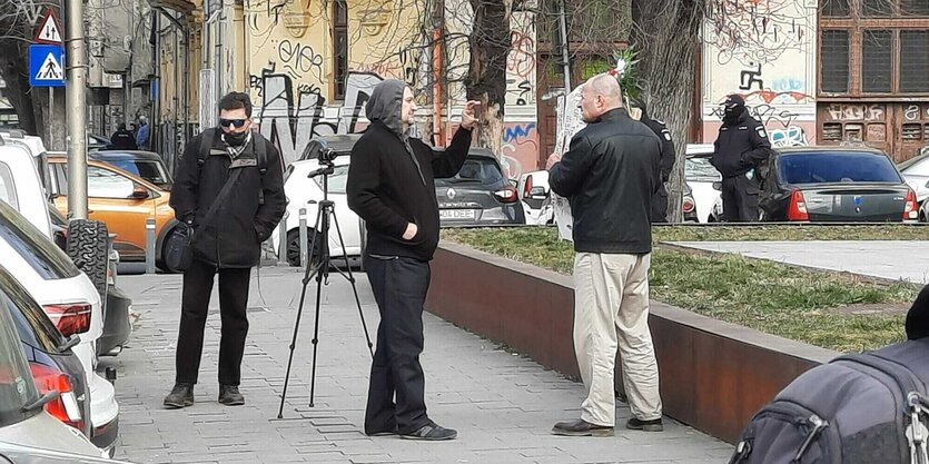 Ein Mann präsentiert einen Kranz, ein zweiter fotografiert mit einem Smartphone