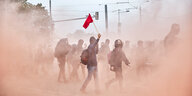 Junge Leute mit OP-Masken gehen durch eine rötliche Rauchwolke, ein junger Mann trägt eine rote Fahne
