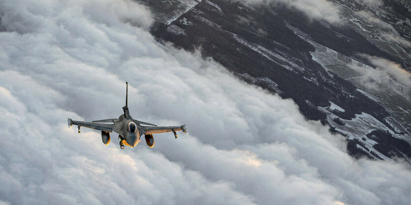 Luftaufnahme eines Kampfflugzeuges, das über eine Wolkendecke fliegt. Weiter im Hintergrund liegt eine Ortschaft