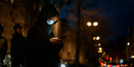 Eine Person mit Kapuze und Maske steht nachts auf der Straße und schaut auf ihr Smartphone