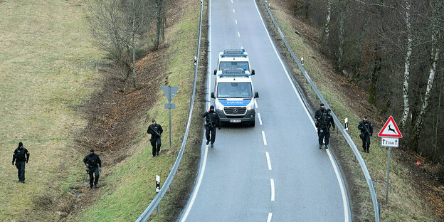 Polizeiwagen auf einer Landstrasse, Polizisten durchkämmen mit Gewehren die Gegend des Tatorts